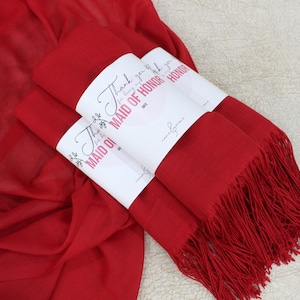 May include: Three red scarves with fringed edges are stacked together. Each scarf has a white label attached with the text "Thank you for being my Maid of Honor".