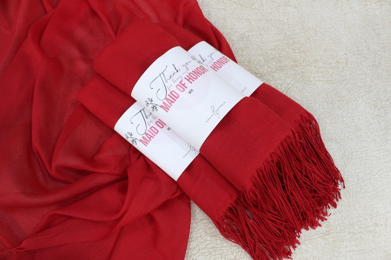 May include: Three red scarves with fringed edges are stacked together. Each scarf has a white label attached with the text "Thank you for being my Maid of Honor".