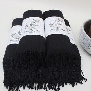 May include: A stack of black pashmina scarves with white labels that say "Stay Warm" and "Cora and John 01/14/2023".