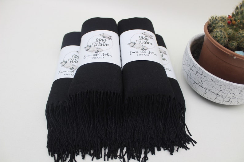 May include: A stack of black pashmina scarves with white labels that say "Stay Warm" and "Cora and John 01/14/2023".