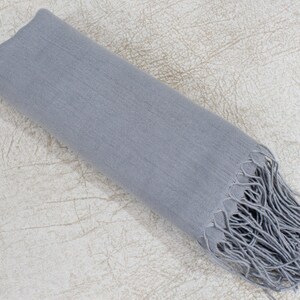 May include: A soft, gray scarf with fringed edges. The scarf is folded in half and laid flat on a textured surface.