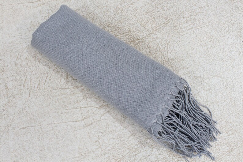May include: A soft, gray scarf with fringed edges. The scarf is folded in half and laid flat on a textured surface.