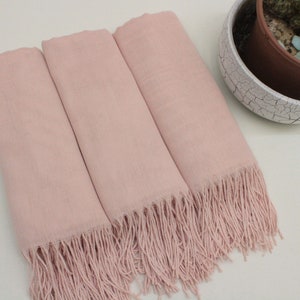 May include: Three pink pashmina scarves with fringe edges. The scarves are folded and arranged on a white surface.