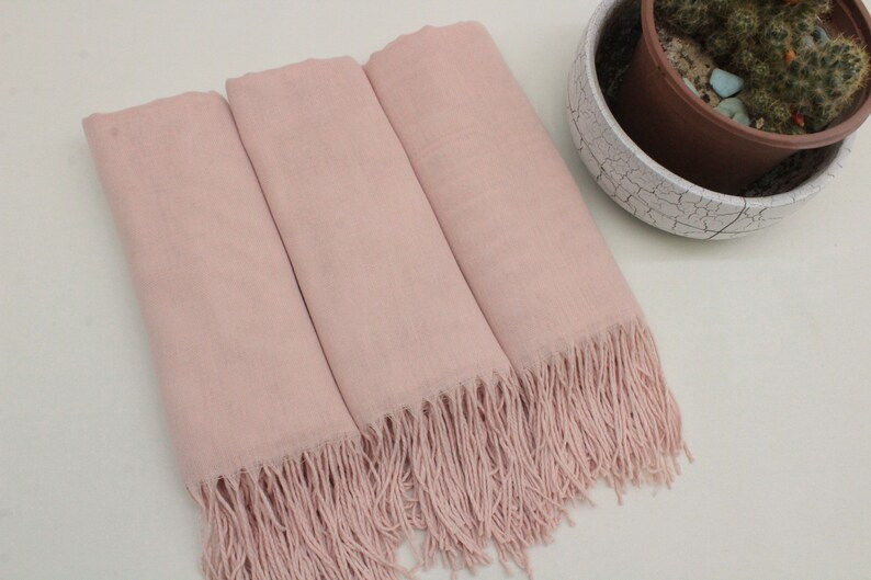 May include: Three pink pashmina scarves with fringe edges. The scarves are folded and arranged on a white surface.