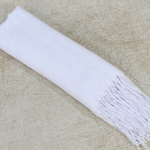 May include: A white scarf with a fringe border. The scarf is folded and laid flat on a textured beige surface.