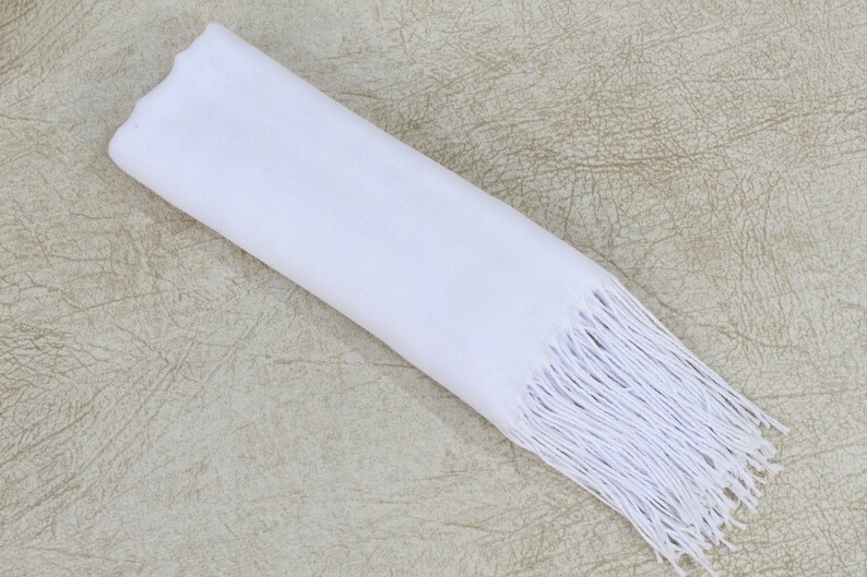 May include: A white scarf with a fringe border. The scarf is folded and laid flat on a textured beige surface.