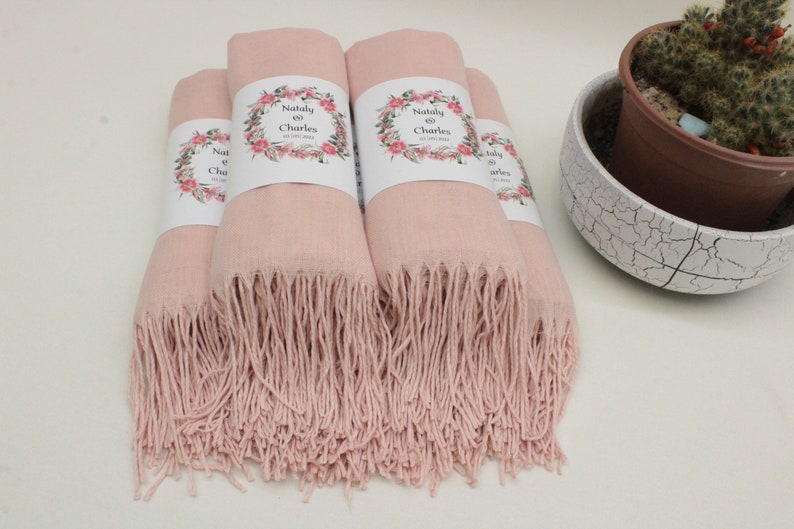 May include: Four pink pashmina scarves with fringe edges. Each scarf has a white label with a floral design and the text "Nataly & Charles" on it.