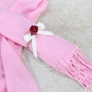 May include: A pink scarf with a white ribbon and a small red rose tied around it. The scarf is rolled up and has fringe on the end.