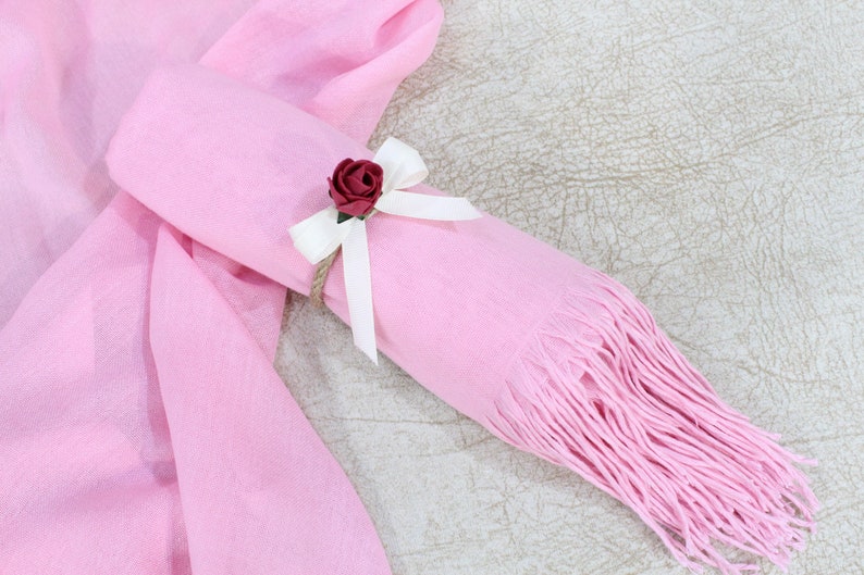 May include: A pink scarf with a white ribbon and a small red rose tied around it. The scarf is rolled up and has fringe on the end.