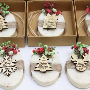 May include: Oval-shaped soaps decorated with dried flowers and wooden ornaments. Each soap is wrapped with twine and placed in a small cardboard box. The ornaments include a Christmas tree, snowflake, gift box, and reindeer, all with "Happy New Year 2024" or "Merry Christmas 2024" text.