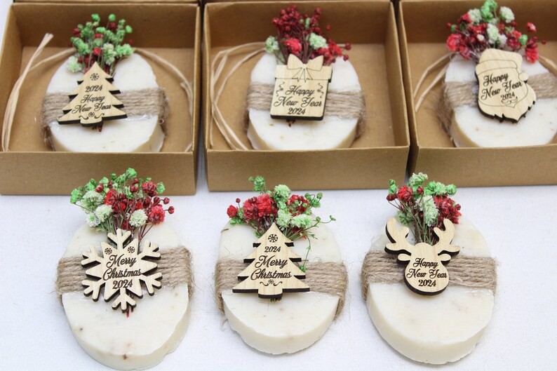May include: Oval-shaped soaps decorated with dried flowers and wooden ornaments. Each soap is wrapped with twine and placed in a small cardboard box. The ornaments include a Christmas tree, snowflake, gift box, and reindeer, all with "Happy New Year 2024" or "Merry Christmas 2024" text.