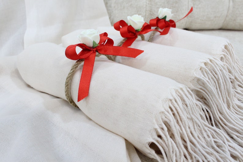 May include: Three white scarves with fringed edges are tied with red ribbon and white flowers.
