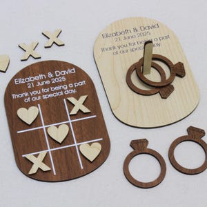 Può includere: Set di giochi di nozze in legno con una scacchiera tris, un gioco di lancio degli anelli e ritagli di anelli con diamanti. La scacchiera tris ha cuori e X. Il gioco di lancio degli anelli ha due anelli. Il testo recita "Elizabeth & David 21 June 2025".