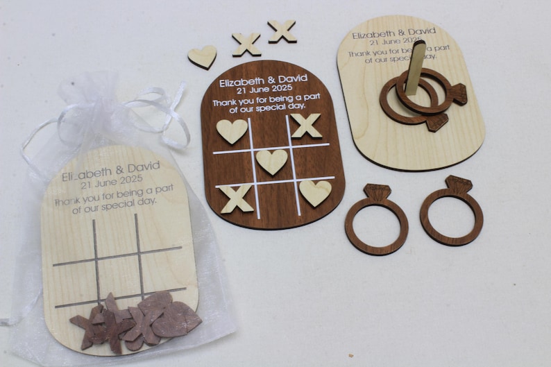 May include: Wooden wedding favors featuring tic-tac-toe games, ring toss, and ring-shaped cutouts. The favors include engraved text with the names "Elizabeth & David" and the date "21 June 2025". The items are in a sheer white bag.