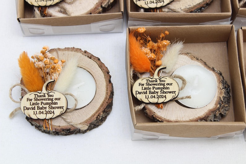 K&ouml;nnte beinhalten: Kerzenhalter aus Holzscheiben mit einem k&uuml;rbisf&ouml;rmigen Anh&auml;nger mit der Aufschrift "Thank You For Showering our Little Pumpkin David Baby Shower 11.04.2024". Jeder Halter hat eine wei&szlig;e Kerze, Trockenblumen und orangefarbene Akzente.