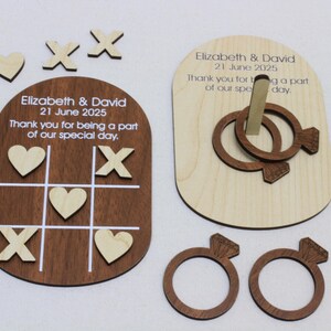 K&ouml;nnte beinhalten: Holz-Hochzeitsspiel-Set mit einem Tic-Tac-Toe-Brett mit Herzen und X's, einem Ringwurfspiel und zwei ringf&ouml;rmigen Spielsteinen. Der Satz enth&auml;lt den Text "Elizabeth & David 21 June 2025".