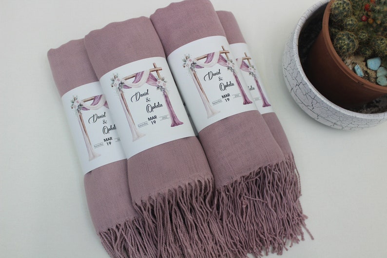 May include: Four dusty rose pashmina shawls with white labels that say "David & Ophelia MAR 19". The labels feature a watercolor illustration of a wedding arch with flowers.