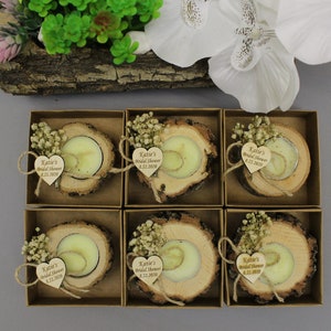 May include: Six small, square boxes each contain a tea light candle set in a rustic wood slice. Each candle is adorned with a small bouquet of dried flowers and a heart-shaped wooden tag with text. The boxes are arranged on a gray surface.