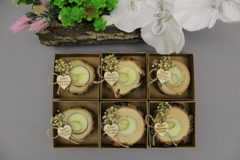 May include: Six small, square boxes each contain a tea light candle set in a rustic wood slice. Each candle is adorned with a small bouquet of dried flowers and a heart-shaped wooden tag with text. The boxes are arranged on a gray surface.