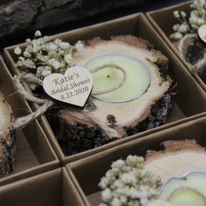 Op de afbeelding: Een set kleine houten kaarshouders met een hartvormig label waarop staat "Katie's Bridal Shower 8.22.2020". Elke houder heeft een kleine witte theelichtkaars en is versierd met gedroogde witte bloemen.