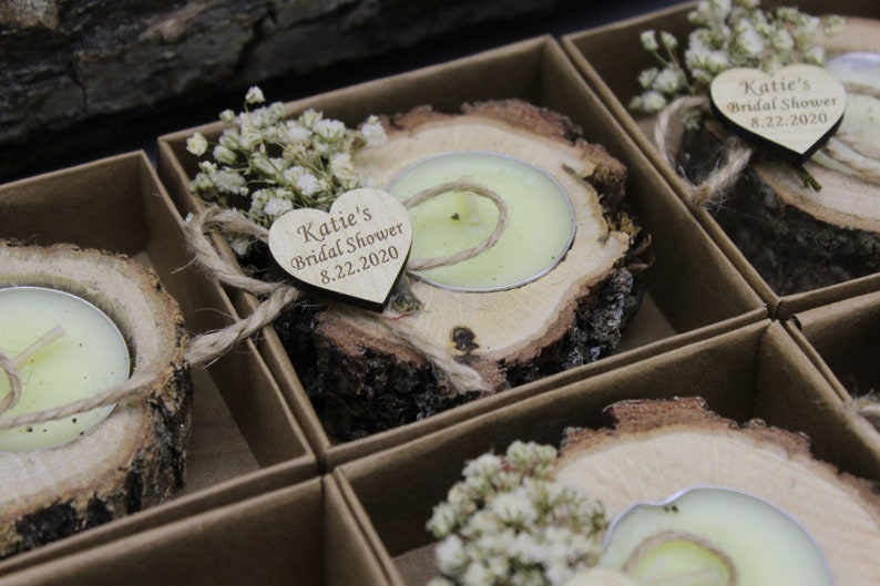 Op de afbeelding: Een set kleine houten kaarshouders met een hartvormig label waarop staat "Katie's Bridal Shower 8.22.2020". Elke houder heeft een kleine witte theelichtkaars en is versierd met gedroogde witte bloemen.