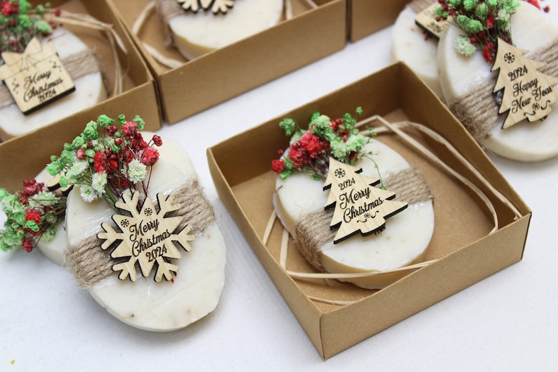 May include: Oval-shaped soaps, wrapped with twine and adorned with dried flowers and wooden ornaments. The ornaments feature the text "Merry Christmas 2024" and snowflake or Christmas tree designs. Presented in small, brown cardboard boxes.