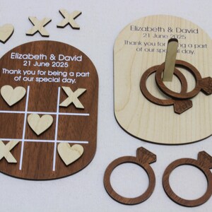 May include: Wooden wedding favors featuring a tic-tac-toe game board and a ring toss game. The tic-tac-toe board is dark brown with white lines and includes wooden hearts and X's. The ring toss game has two interlocking rings. Text includes names and a date.