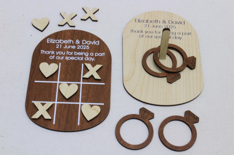 K&ouml;nnte beinhalten: Hochzeitsgeschenke aus Holz mit einem Tic-Tac-Toe-Spielbrett und einem Ringwurfspiel. Das Tic-Tac-Toe-Spielbrett ist dunkelbraun mit wei&szlig;en Linien und enth&auml;lt Holzherzen und X's. Das Ringwurfspiel hat zwei ineinandergreifende Ringe. Der Text enth&auml;lt Namen und ein Datum.