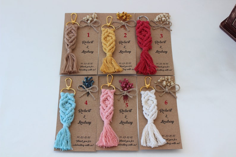Puede incluir: Seis llaveros con un dise&ntilde;o de macram&eacute; en diferentes colores, incluyendo beige, amarillo, rojo, azul, rosa y blanco. Cada llavero tiene un anillo dorado y una borla. Los llaveros est&aacute;n unidos a peque&ntilde;as tarjetas con el texto "Robert & Lindsay" y la fecha "10.23.2020".