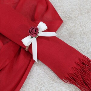 May include: A red scarf with a white bow and a small red rose tied around it. The scarf has a fringe on the end.
