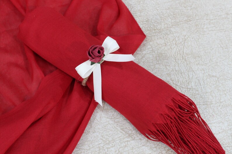 May include: A red scarf with a white bow and a small red rose tied around it. The scarf has a fringe on the end.