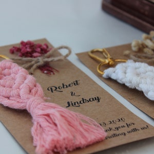 Puede incluir: Dos llaveros de recuerdo de boda con un dise&ntilde;o de macram&eacute; rosa y blanco. Los llaveros est&aacute;n unidos a tarjetas marrones con el texto "Robert & Lindsay" y "&iexcl;Gracias por celebrar con nosotros!"