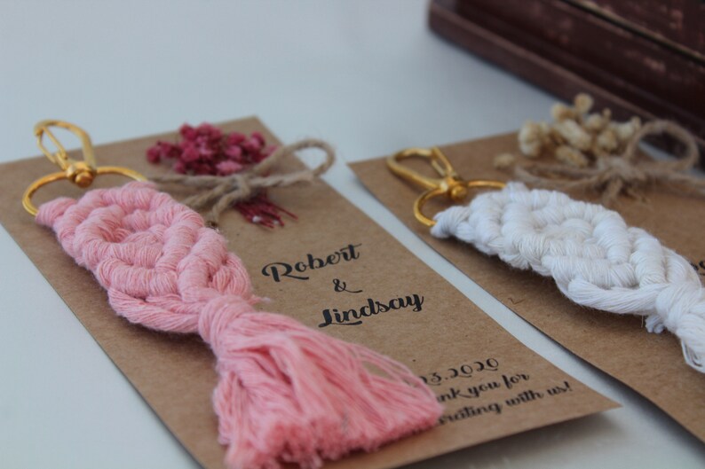 Puede incluir: Dos llaveros de recuerdo de boda con un dise&ntilde;o de macram&eacute; rosa y blanco. Los llaveros est&aacute;n unidos a tarjetas marrones con el texto "Robert & Lindsay" y "&iexcl;Gracias por celebrar con nosotros!"