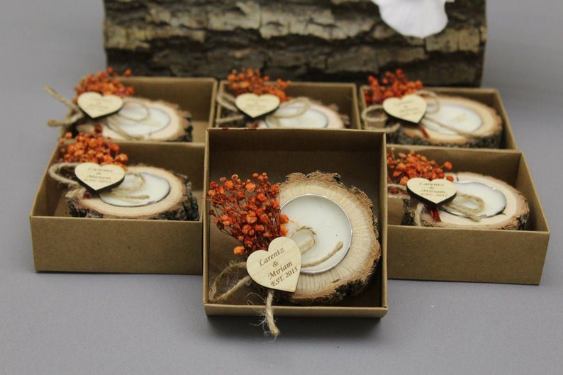 Op de afbeelding: Set van kleine, rustieke houten kaarshouders met een kleine witte kaars in elk. Elke houder is versierd met een klein hartvormig houten label met de tekst "Lauren & Miriam Est. 2021" en een klein bosje gedroogde oranje bloemen.