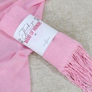 May include: A pink scarf with fringe is rolled up and tied with a white tag that says "Thank you for being my Maid of Honor".