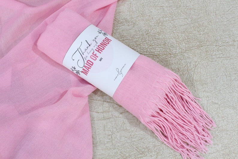 May include: A pink scarf with fringe is rolled up and tied with a white tag that says "Thank you for being my Maid of Honor".
