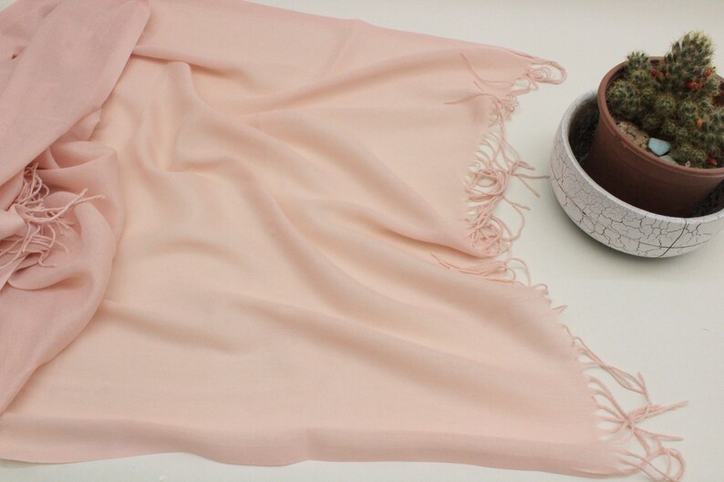 May include: A soft, pale pink scarf with a fringed edge. The scarf is made of a lightweight fabric and is perfect for adding a touch of color to any outfit.