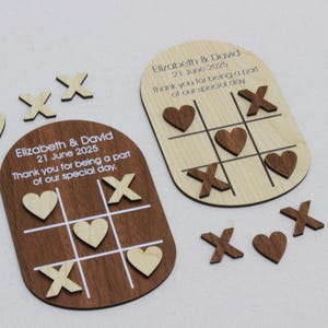 May include: Wooden tic-tac-toe game pieces and boards. One board is dark brown, the other is light wood. The boards have the names "Elizabeth & David" and the date "21 June 2025" printed on them. The game pieces include hearts and X's.