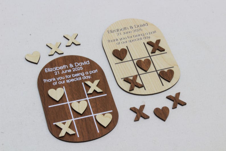 May include: Wooden tic-tac-toe game pieces and boards. One board is dark brown, the other is light wood. The boards have the names "Elizabeth & David" and the date "21 June 2025" printed on them. The game pieces include hearts and X's.