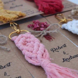 Puede incluir: Llaveros de macram&eacute; rosa, blanco y amarillo con flores secas y un anillo de llave dorado. Los llaveros est&aacute;n unidos a tarjetas con el texto "Robert & Lindsay" y la fecha "25.5.2020".