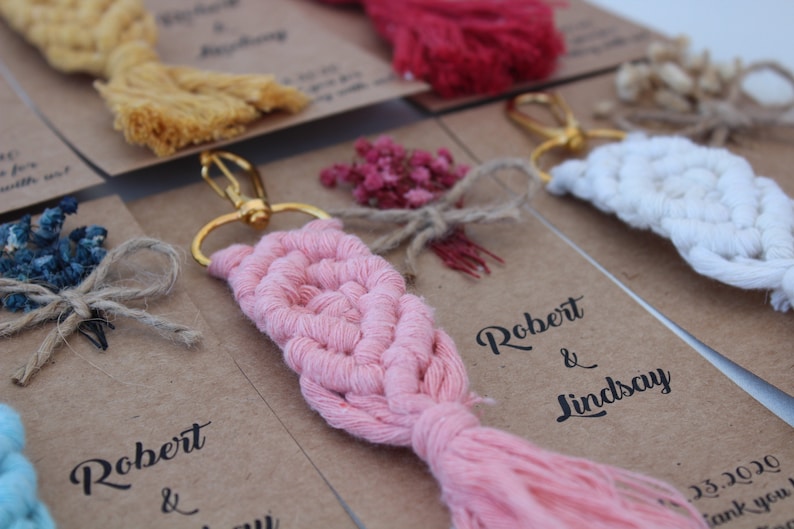 Puede incluir: Llaveros de macram&eacute; rosa, blanco y amarillo con flores secas y un anillo de llave dorado. Los llaveros est&aacute;n unidos a tarjetas con el texto "Robert & Lindsay" y la fecha "25.5.2020".