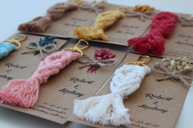 Puede incluir: Un conjunto de seis llaveros con una variedad de colores y texturas. Cada llavero presenta un borla trenzada con un anillo de llave dorado. Los llaveros se muestran en tarjetas marrones con el texto "Robert & Lindsay" y la fecha "11.23.2020".