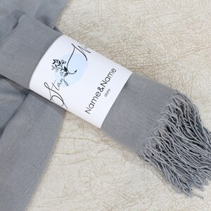May include: A gray pashmina scarf with fringe, rolled up and tied with a white label that says "Stay" and "Name & Name date".