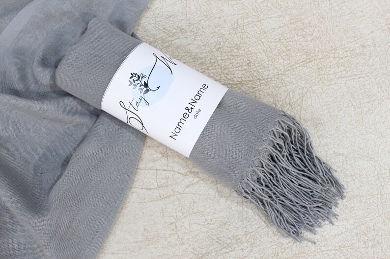 May include: A gray pashmina scarf with fringe, rolled up and tied with a white label that says "Stay" and "Name & Name date".
