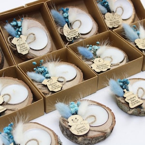 Op de afbeelding: Een verzameling kleine, rustieke houten kaarshouders met gedroogde bloemen en gepersonaliseerde labels. Elke houder heeft een klein theelichtje en een houten label met een naam en eventdetails, zoals "Baby Shower" of "Doop".