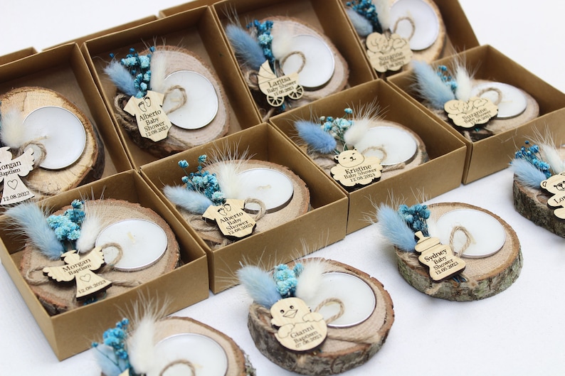 Op de afbeelding: Een verzameling kleine, rustieke houten kaarshouders met gedroogde bloemen en gepersonaliseerde labels. Elke houder heeft een klein theelichtje en een houten label met een naam en eventdetails, zoals "Baby Shower" of "Doop".