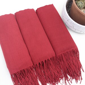 May include: Three burgundy pashmina scarves with fringe. The scarves are folded and stacked on top of each other.