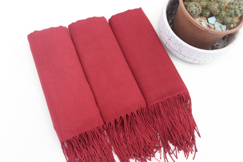 May include: Three burgundy pashmina scarves with fringe. The scarves are folded and stacked on top of each other.