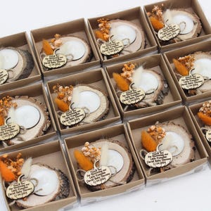 K&ouml;nnte beinhalten: Babyparty-Gastgeschenke in kleinen Pappschachteln. Jede Schachtel enth&auml;lt einen h&ouml;lzernen Kerzenhalter mit einer wei&szlig;en Kerze, Trockenblumen und einem Holzanh&auml;nger mit der Aufschrift "Thank You for Showering our Little Pumpkin David Baby Shower 12.04.2024."