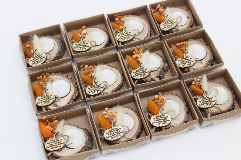 K&ouml;nnte beinhalten: Babyparty-Gastgeschenke in kleinen Pappschachteln. Jede Schachtel enth&auml;lt einen h&ouml;lzernen Kerzenhalter mit einer wei&szlig;en Kerze, Trockenblumen und einem Holzanh&auml;nger mit der Aufschrift "Thank You for Showering our Little Pumpkin David Baby Shower 12.04.2024."